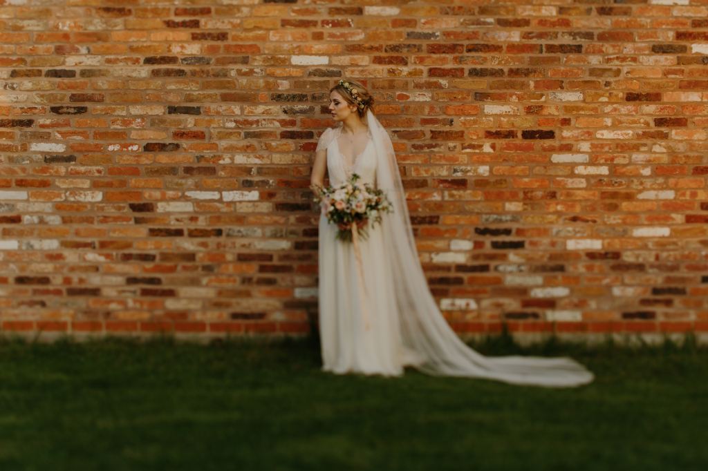A bride standing in-front of a brick wall, holding her bouquet and head turned to the left, her eyes are closed.